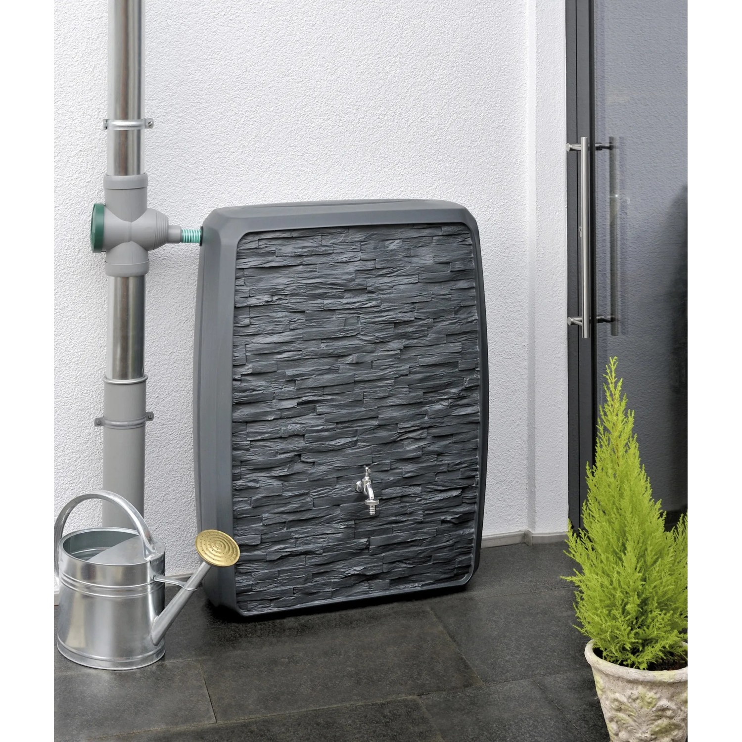 Anthrazitfarbene Waterform Regentonne Multitank 250 l mit Steinoptik und Wasserhahn neben Gießkanne.