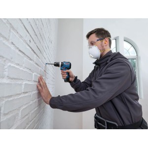 Mann bohrt mit Bosch PRO Akku-Schlagbohrmaschine GSB12V-31 in weiße Ziegelwand. Trägt Schutzbrille und Staubmaske.