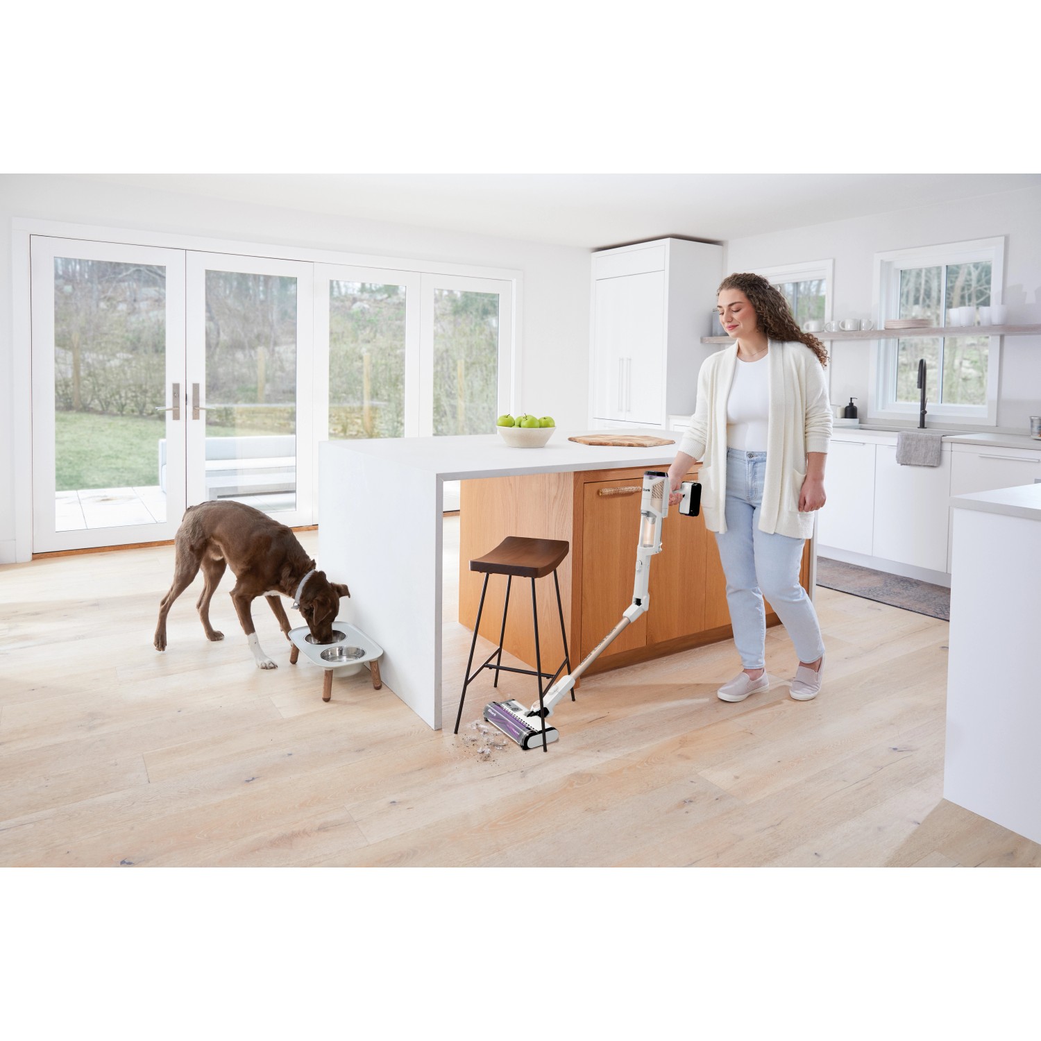 Frau nutzt Shark Akkusauger Detect Pro mit Absaugstation in Küche, Hund frisst. Bodenstaubsauger für Tierhaare.