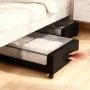Schwarze Unterbettkommode mit transparentem Deckel und Rollen, gefüllt mit Kleidung, wird unter Bett hervorgezogen. Praktischer Stauraum.