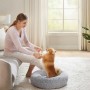 Frau spielt mit kleinem Hund in grauem, flauschigem Hundebett. Das runde Hundesofa bietet Komfort.
