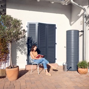 Garantia NORDIC Regenspeicher 500 l in Graphite Grey neben Hauswand mit Frau lesend im Garten.