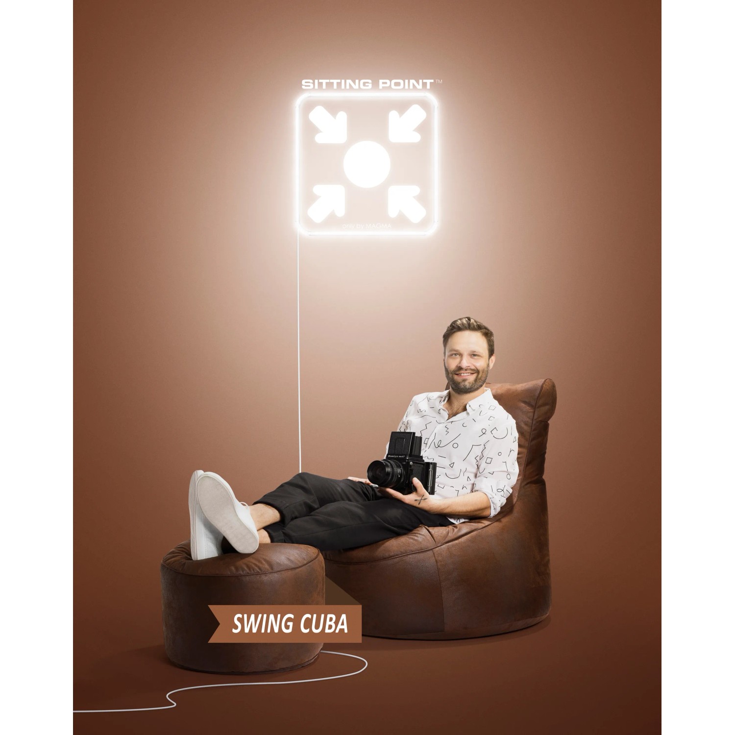 Mann entspannt auf braunem Sitting Point Hocker/Sitzsack mit Kamera. Bequemer Hocker mit Seitentasche.