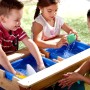 Kinder spielen am Axi Sand- und Wasser-Picknicktisch Nick mit blauen Wannen und Sonnenschirm.