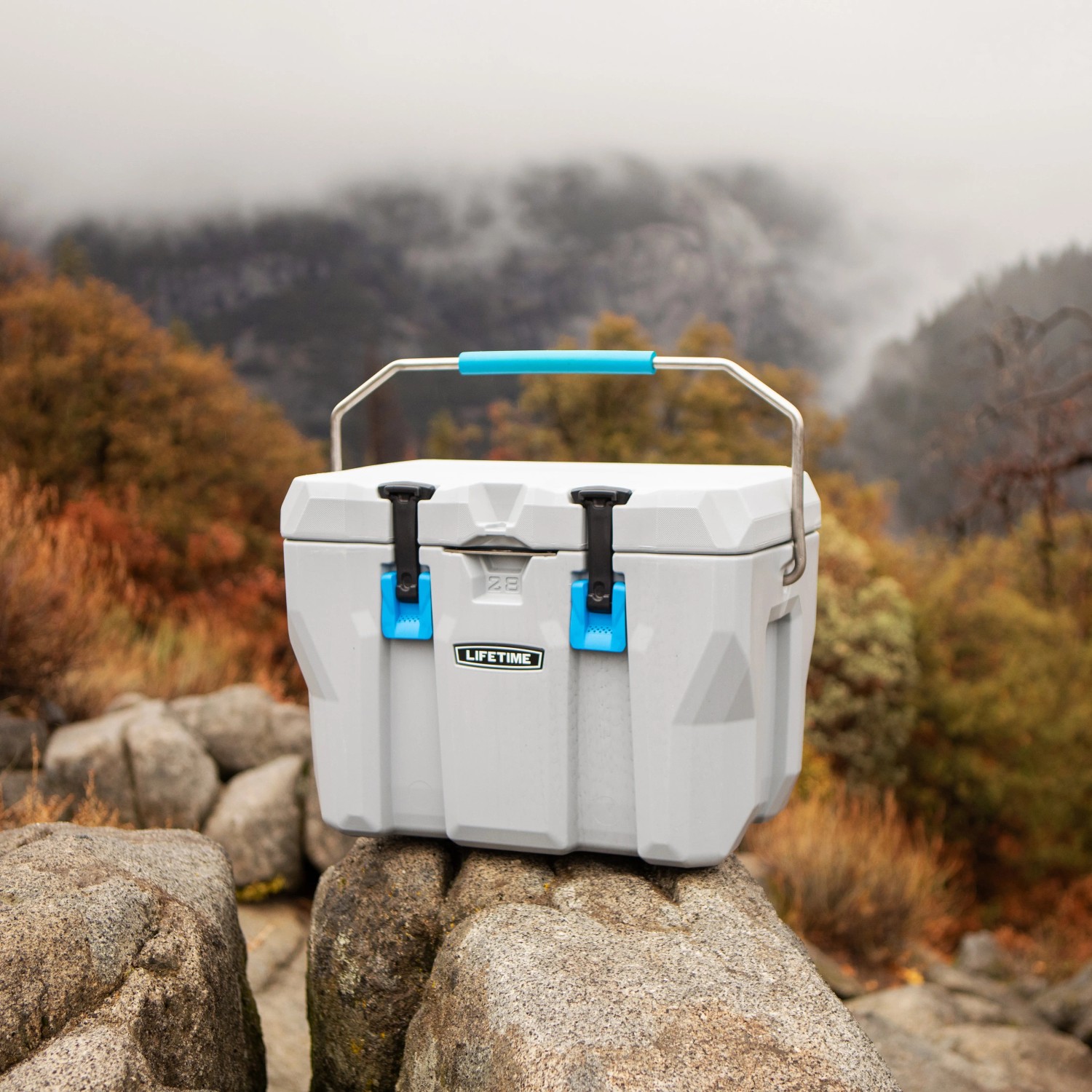 Graue Lifetime Kühlbox Campingbox (26,5 Liter) mit blauem Griff auf Felsen.