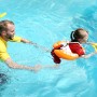 Kind mit gelbem Freds Swim Academy Schwimmreifen Classic im Pool, betreut von einem Erwachsenen.