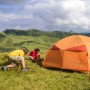 Zwei Personen befestigen ein orangefarbenes Zelt mit Felsbodenheringen in bergiger Landschaft. Perfekt für Camping.