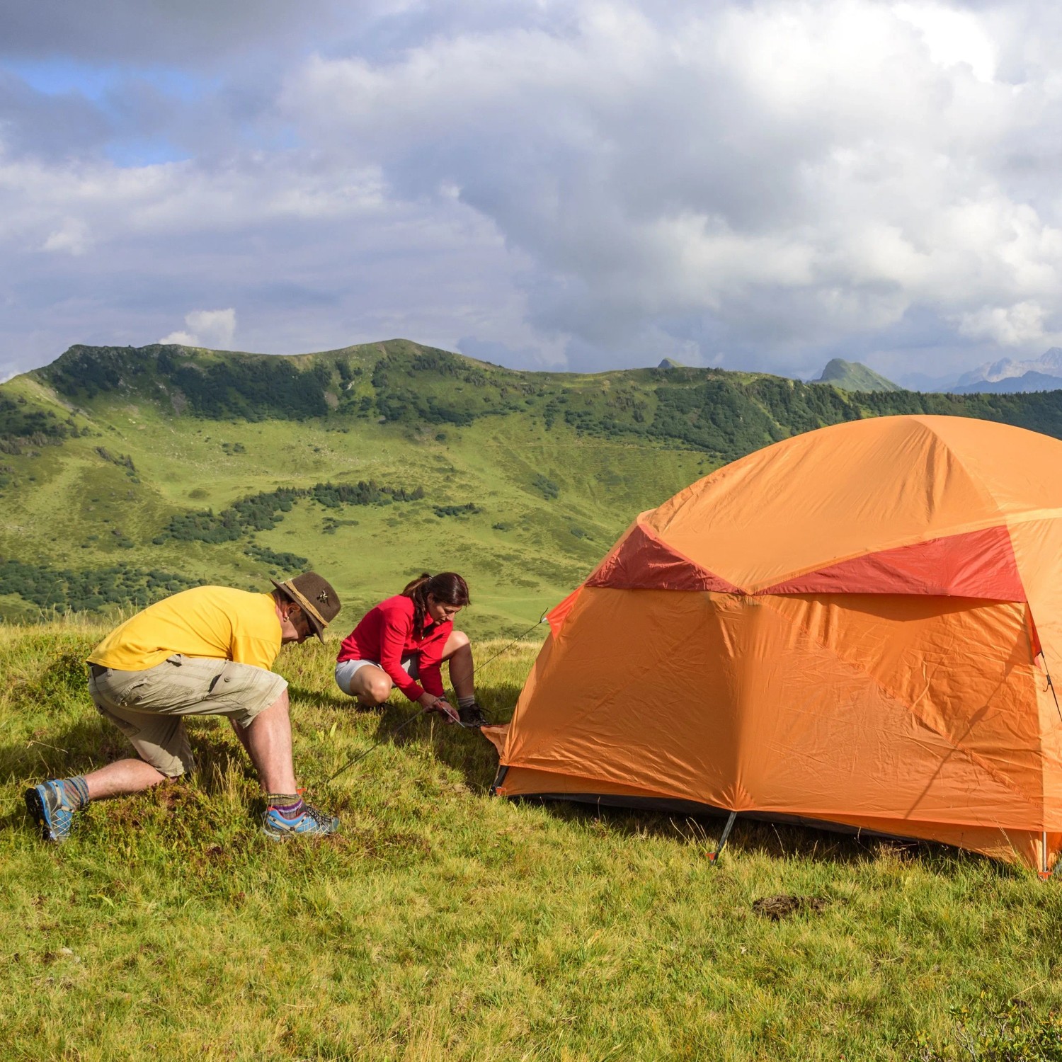 Zwei Personen befestigen ein orangefarbenes Zelt mit Felsbodenheringen in bergiger Landschaft. Perfekt für Camping.