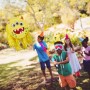 Gelbe Monster Pinata aus Pappe mit drei Augen hängt im Garten. Kinder spielen blindlings das Party-Spiel. Perfekt für Kindergeburtstage.