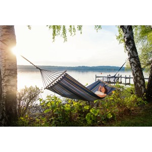 Frau entspannt in der Stabhängematte Tonga Ocean (200x100 cm) zwischen zwei Bäumen am See.