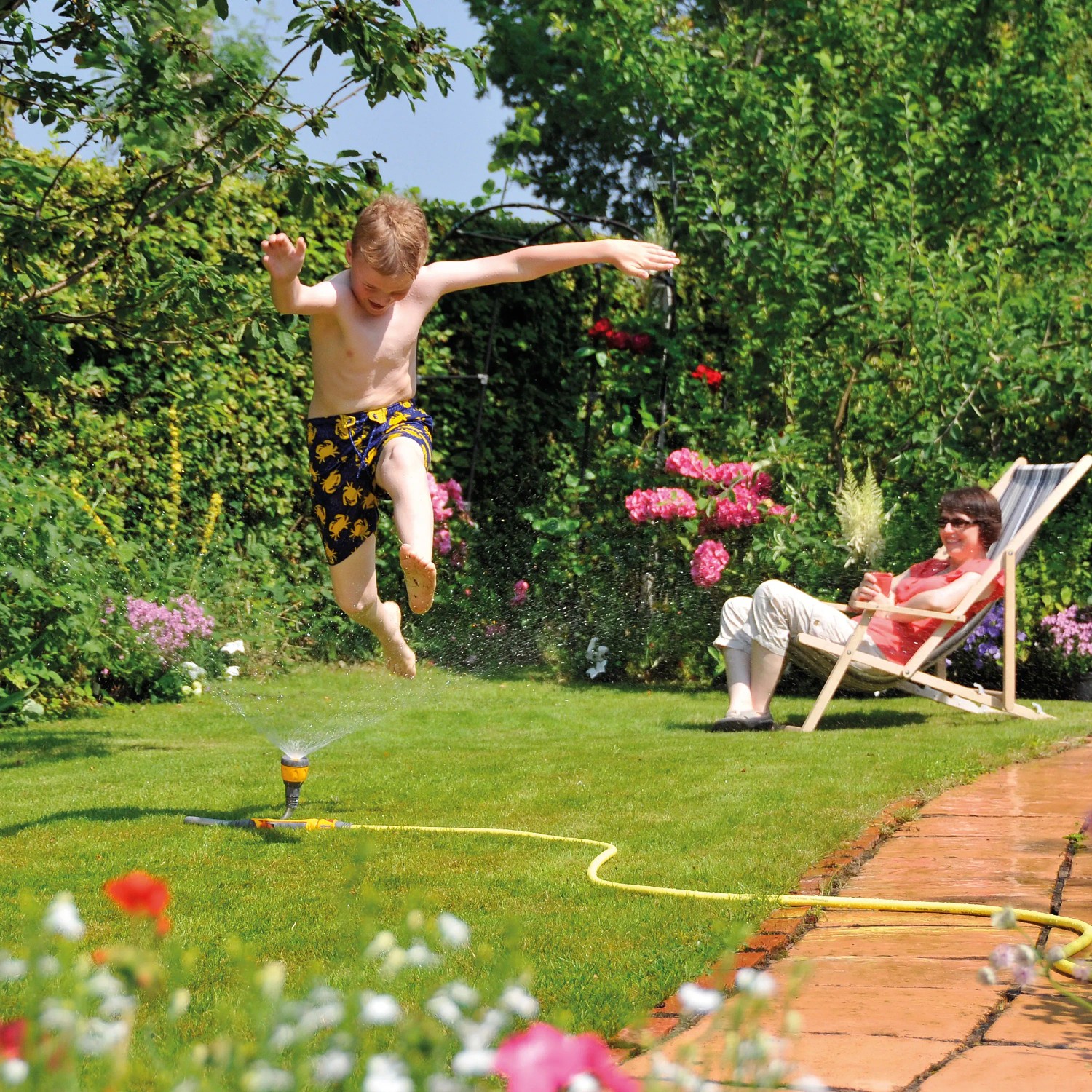 Kind springt über Hozelock Flexi Spray Gartenbrause, während Frau im Liegestuhl entspannt.