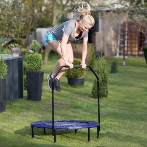 Frau springt auf blauem Trampolin mit schwarzer Haltestange. Fitnessgerät für Balance, Kondition & Koordination.