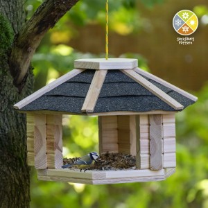 Dobar Vogelhaus aus hellem Massivholz mit schwarzem Dach, hängt im Garten. Eine Meise frisst im Futtertablett.