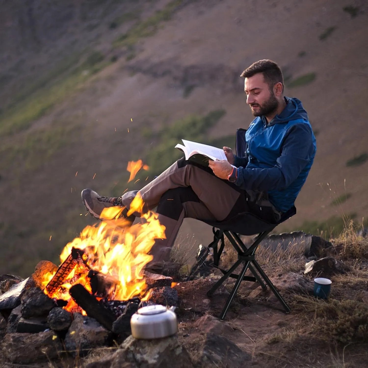 Mann liest auf schwarzem Klapphocker am Lagerfeuer. Faltbarer Camping Dreibeinhocker für Outdoor-Aktivitäten.