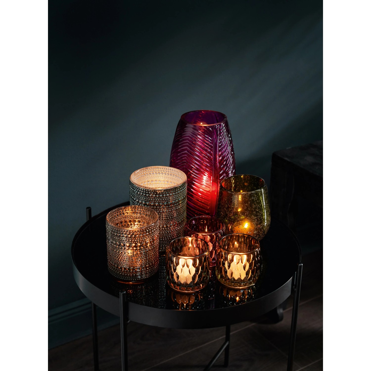 Verschiedene Teelichthalter der Serie Dark Wonder aus Glas in Grün-, Gold- und Lilatönen auf einem Tisch.