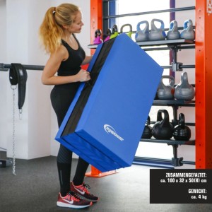 Frau trägt gefaltete blaue eyepower Weichbodenmatte mit Tragegriff im Fitnessraum. Ideal für Gymnastik & Sport.