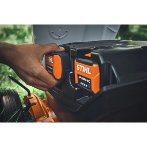 Hand setzt orange-schwarzen Stihl AP Akku in den Akku-Rasenmäher Stihl RMA 7 RV ein. Zwei Akkus sichtbar.