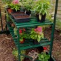 Grüner Alutisch (121x54cm) mit 2 Ablagen im Gewächshaus, bepflanzt mit Blumen.
