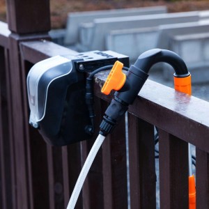Yard Force Akku-Regenfasspumpe: Batteriekasten mit Schlauchanschluss und orangefarbenem Ventil an Holzgeländer.