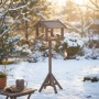 Braunes Relaxdays Vogelfutterhaus mit Ständer im winterlichen Garten mit Vögeln.