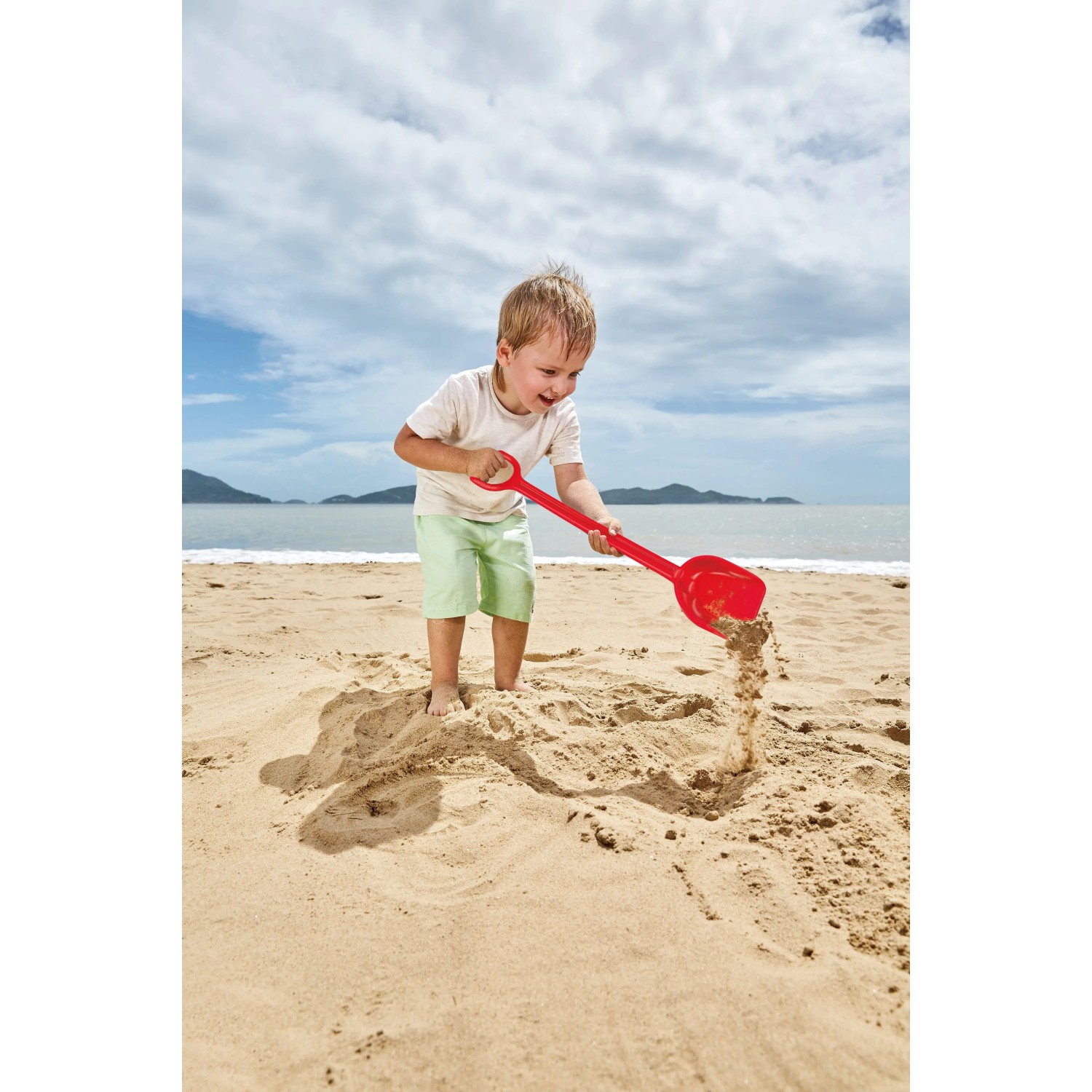 Kind spielt mit roter Hape Schaufel (40 cm) im Sand am Strand.