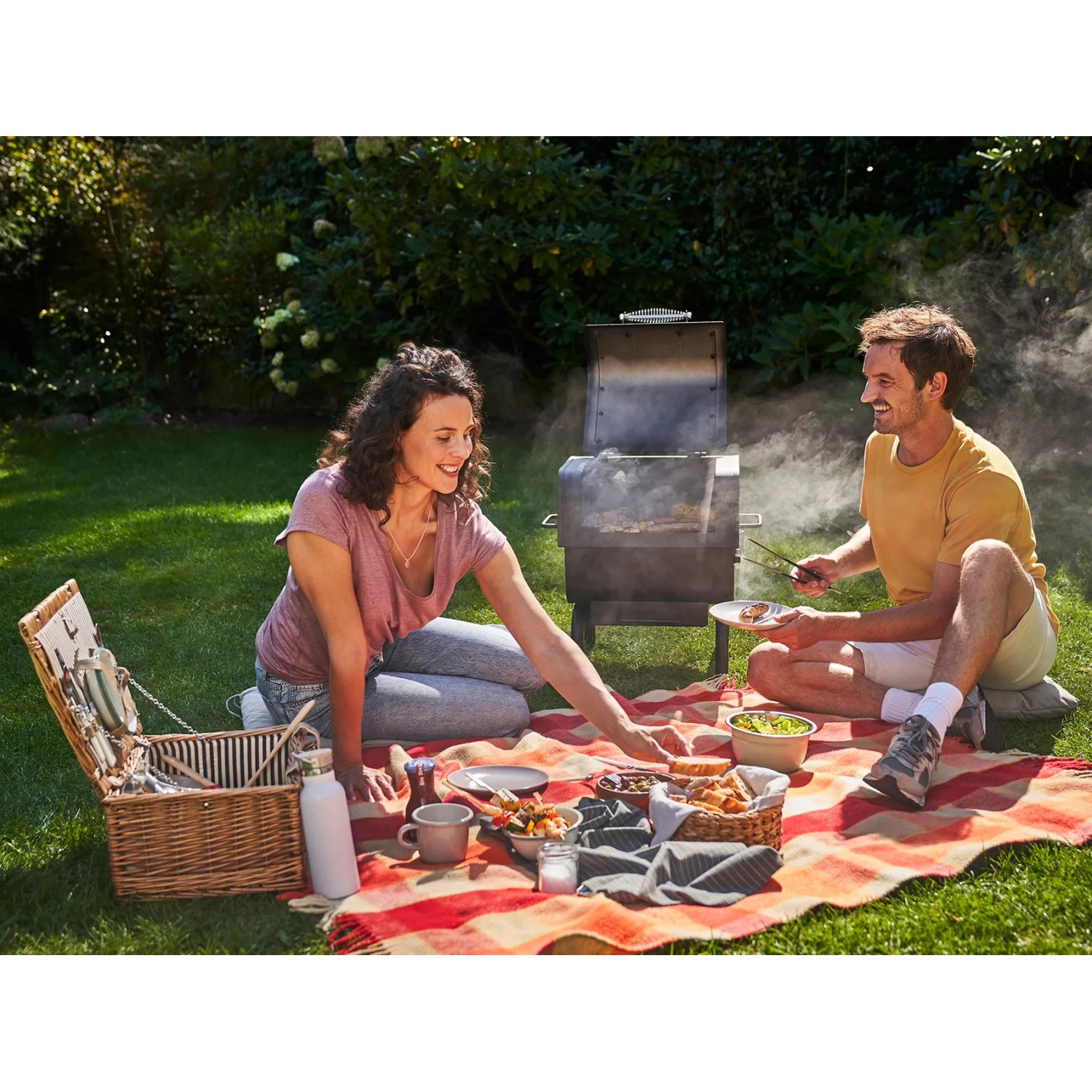 Paar grillt mit dem tragbaren Char-Broil Holzkohlegrill 2Go im Garten beim Picknick.