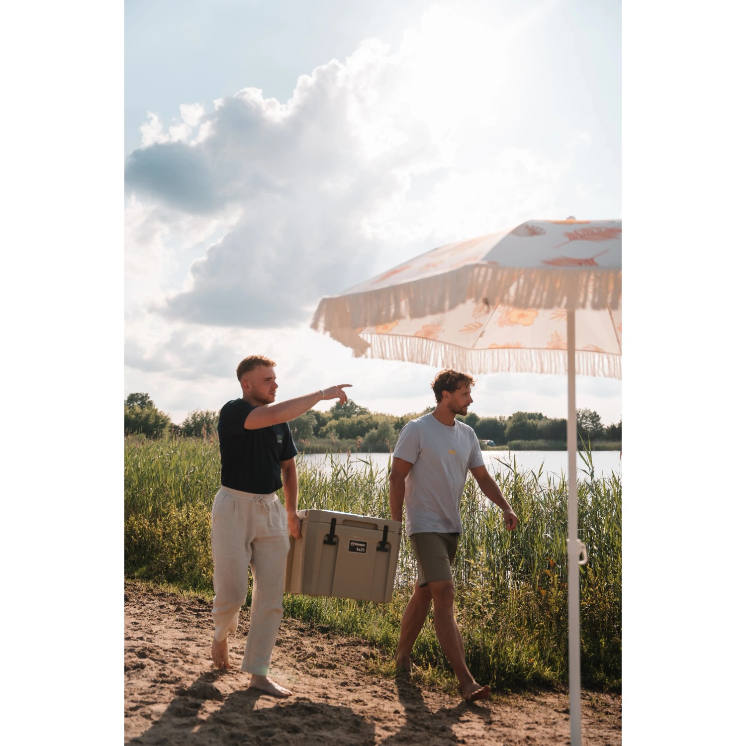 Zwei Personen tragen eine sandfarbene Petromax Kühlbox 25l am See.
