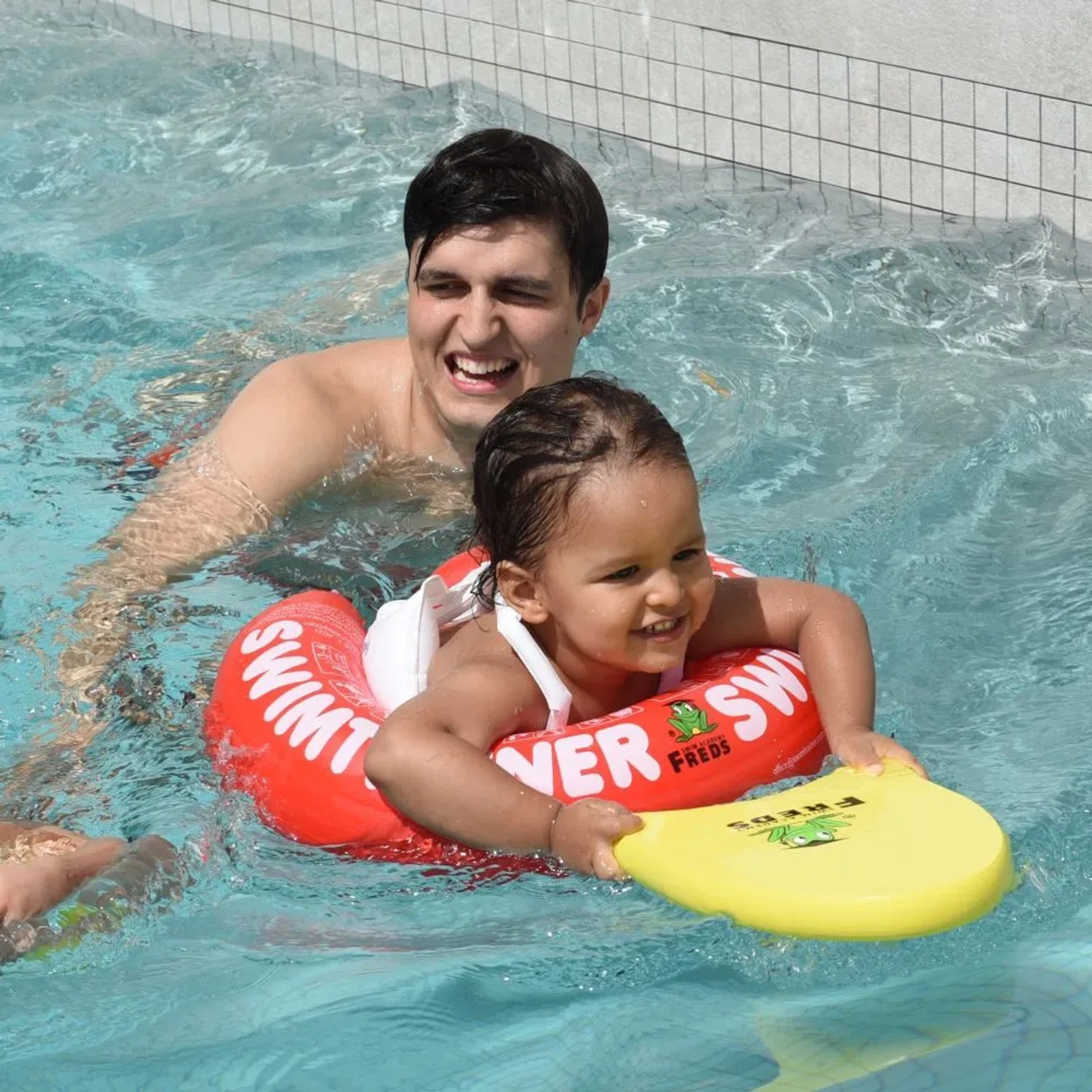 Kind mit rotem Freds Swim Academy Schwimmreifen Classic im Pool, begleitet von einem Erwachsenen.