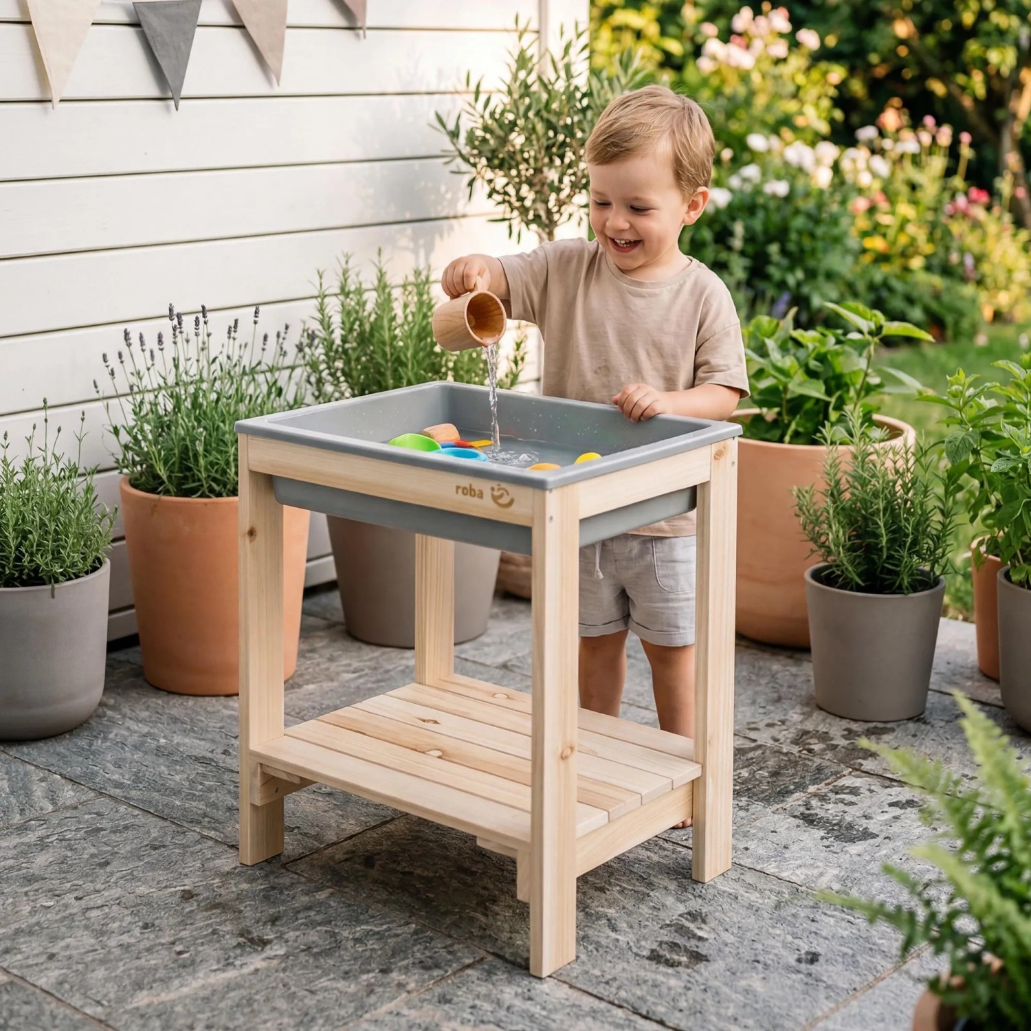 Lachendes Kind spielt mit Wasser am roba Outdoor Spiel- & Wassertisch aus Naturholz mit grauer Wanne auf der Terrasse.
