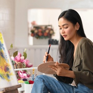 Frau malt Bild mit Pinsel und Palette, Rollwagen im Hintergrund