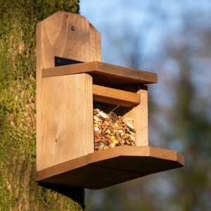 Braune Eichhörnchen-Futterstation aus Holz, gefüllt mit Futter, an einem Baumstamm. Robustes Futterhaus für Eichhörnchen.