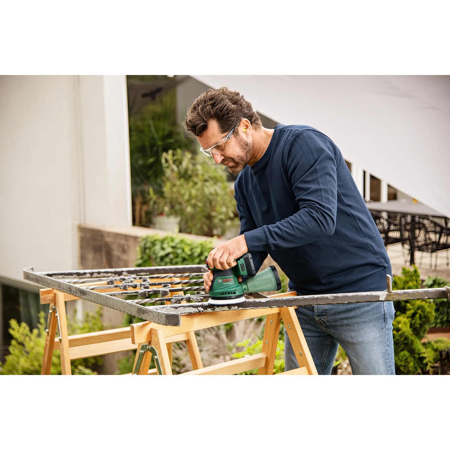 Mann schleift Metall mit grünem Bosch Akku-Exzenterschleifer UniversalOrbit 18V-20. Schleifmaschine für Heimwerker.