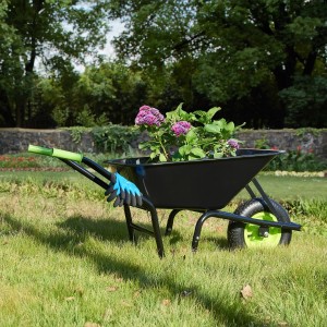 Schwarze Juskys Schubkarre Garden 100L mit grünen Griffen und blauen Gartenhandschuhen im Garten.