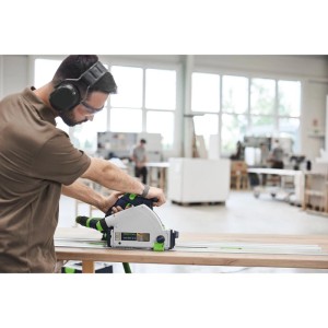 Mann sägt mit Festool Akku-Tauchsäge TSC55 KSEB-Basic präzise Holzplatte. Graue Säge mit grünem Akku auf Führungsschiene in Werkstatt.