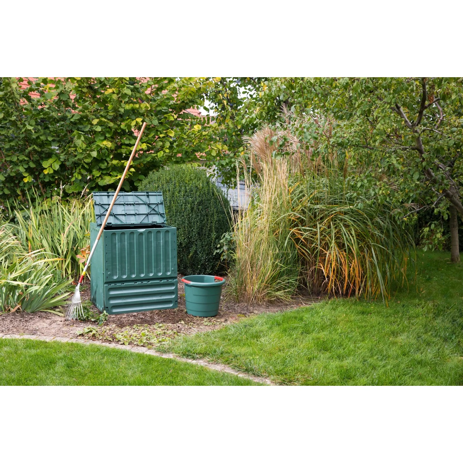Grüner Garantia Komposter Comfort Eco-King 600 l im Garten mit Laubrechen und Eimer.