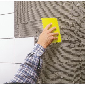 Gelber LUX Spachtelsatz Classic beim Auftragen von Kleber auf Fliesenwand.