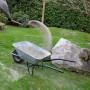 Person schaufelt hellgrauen Galamio Rasensand in Schubkarre; Garten mit Gras, Buschwerk.