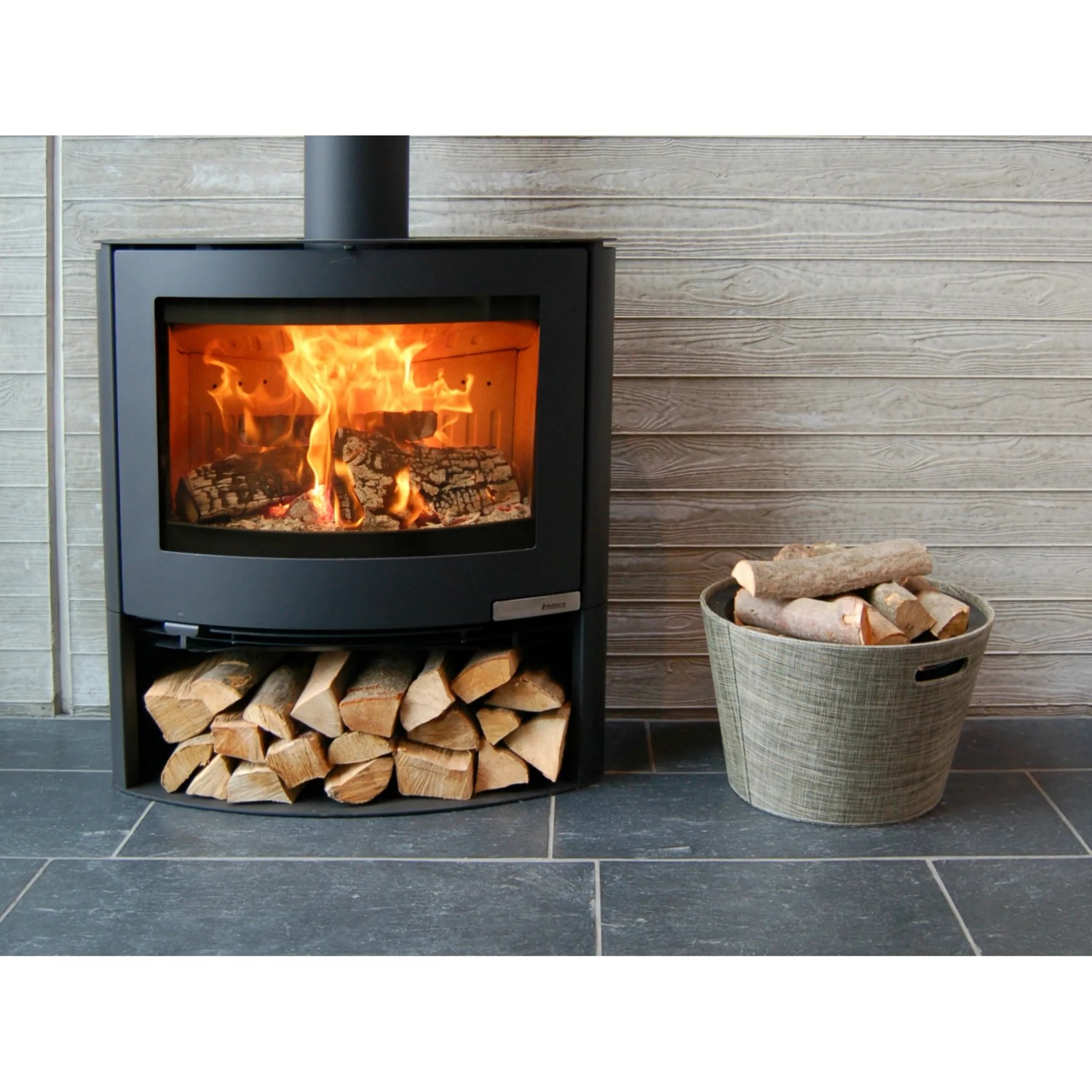 Schwarzer Aduro Kaminofen 15.2 mit Holzscheiten und Korb. Der Kaminofen hat eine Automatik und 6,5 kW Leistung.