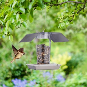 Silbernes Edelstahl Vogelfutterhaus mit transparentem Silo, gefüllt mit Futter, hängt im Garten. Ein Wildvogel fliegt heran.