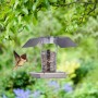 Silbernes Edelstahl Vogelfutterhaus mit transparentem Silo, gefüllt mit Futter, hängt im Garten. Ein Wildvogel fliegt heran.