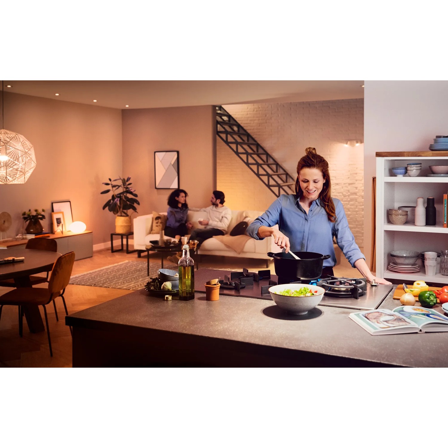 Szene mit Philips Hue White Ambiance E27 LED: Frau kocht, Paar entspannt im Wohnzimmer, warmes Licht.