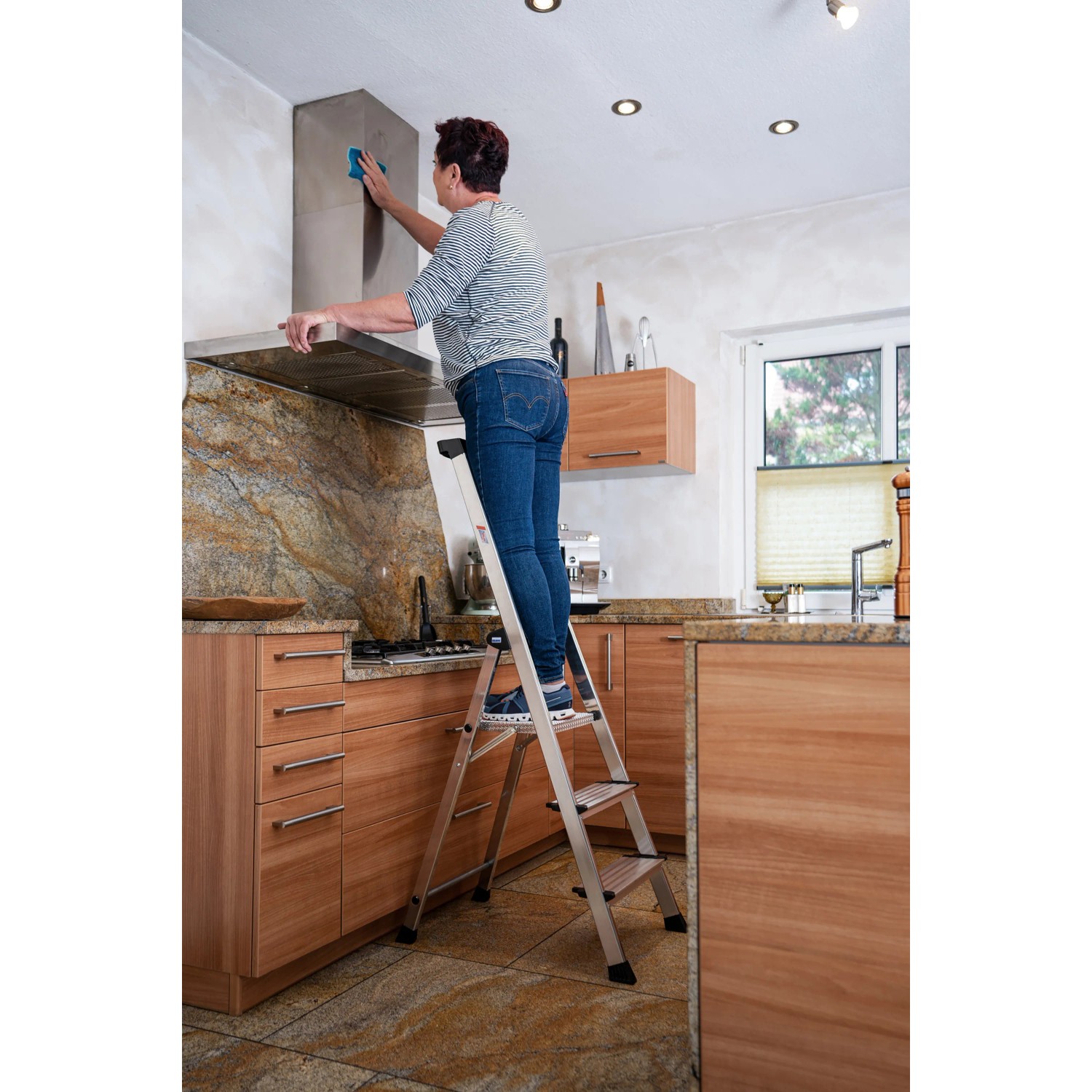 Frau auf Krause Stehleiter Safety Plusline (3 Stufen) in Küche, beim Putzen der Dunstabzugshaube.