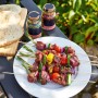 Grillspieße mit Gemüse und Fleisch auf einem Teller, Brot und Gewürze im Hintergrund.