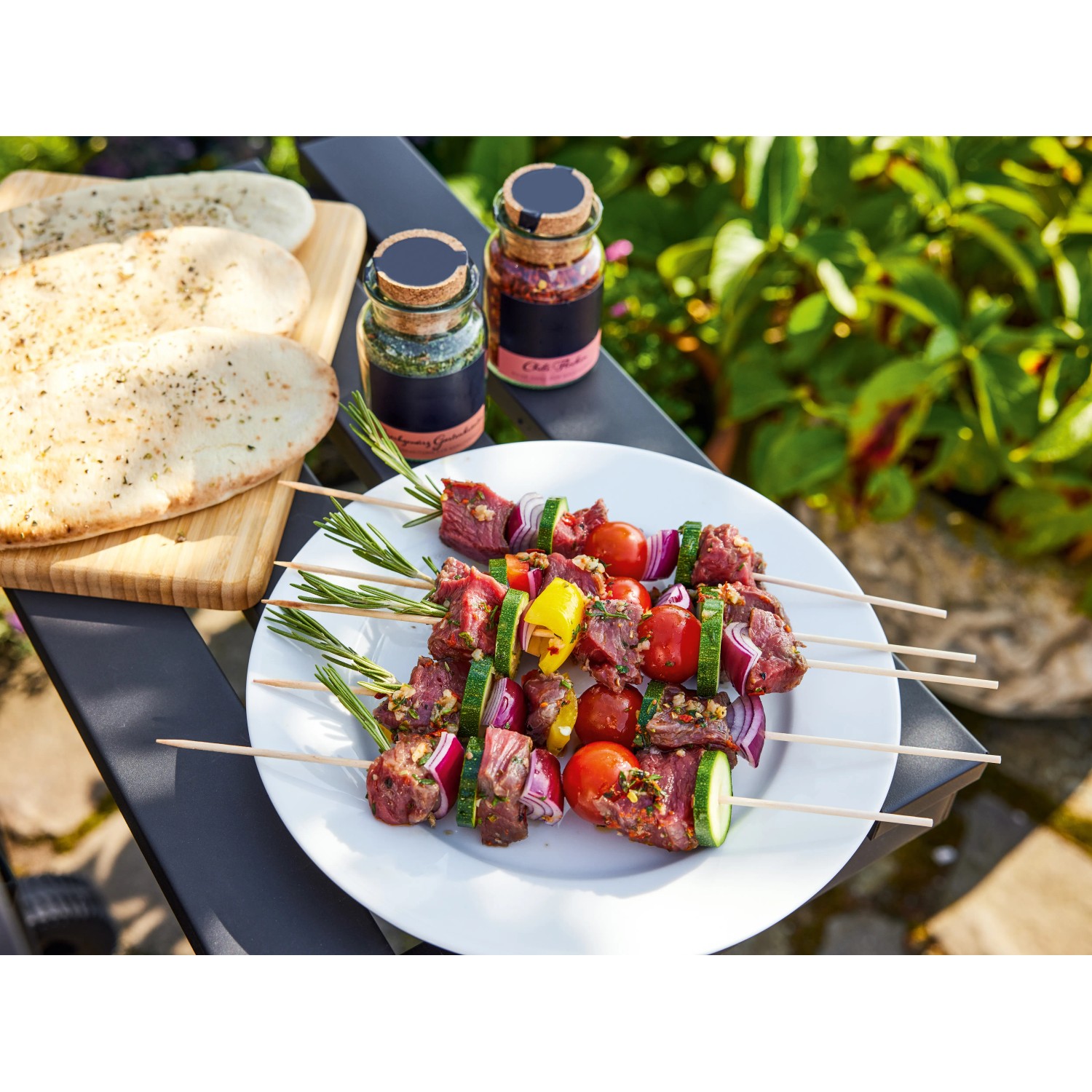 Grillspieße mit Gemüse und Fleisch auf einem Teller, Brot und Gewürze im Hintergrund.