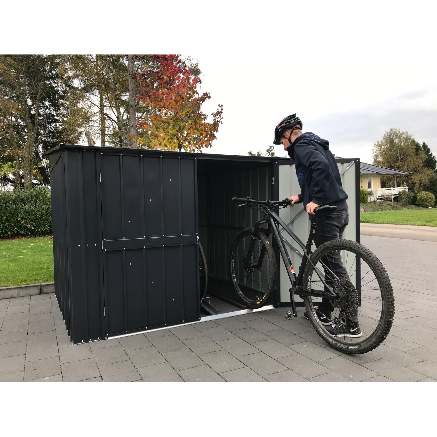 Anthrazitfarbene Globel Fahrradbox Family mit 4 Fahrradhaltern, mit Person und Fahrrad vor der offenen Box.