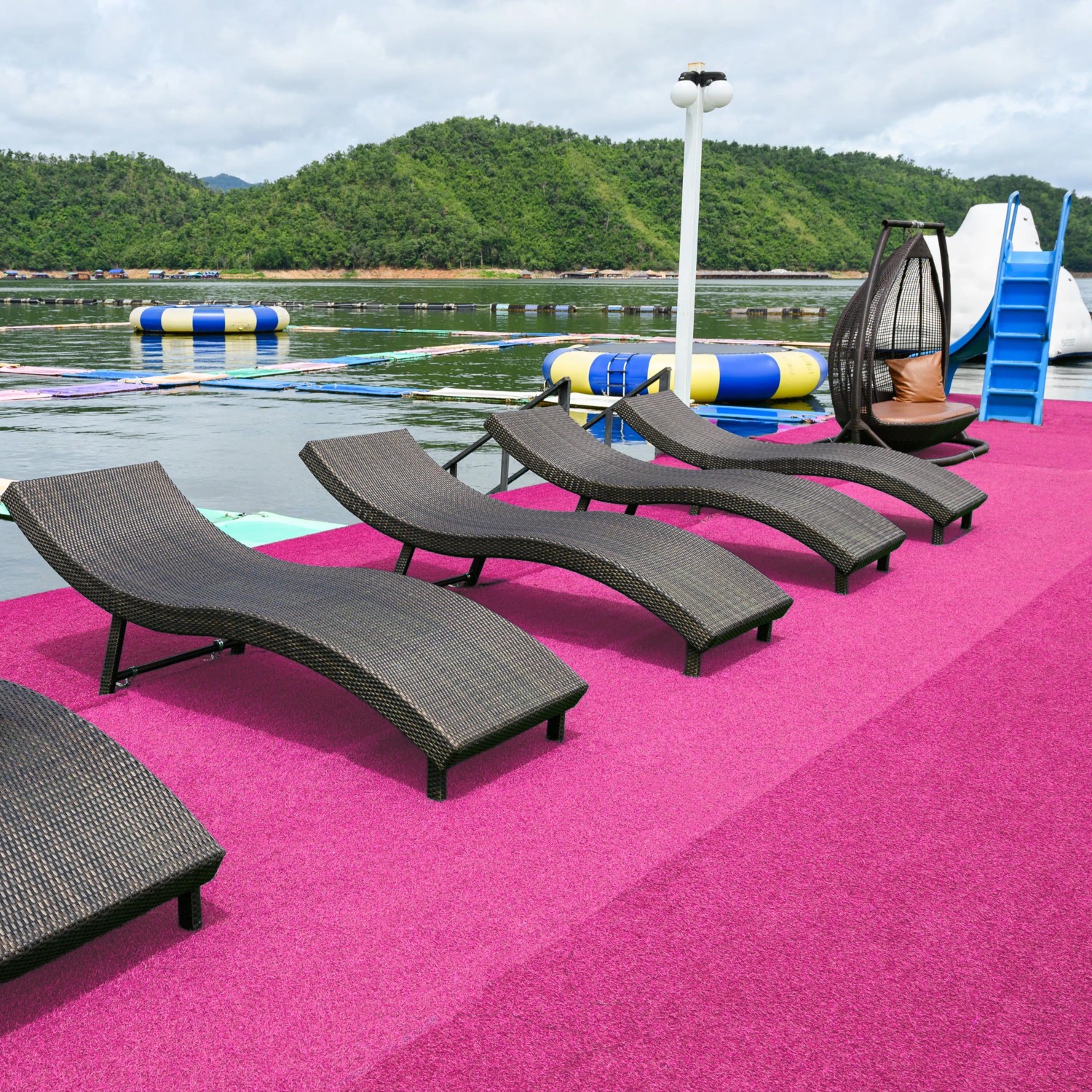 Pinker Primaflor Kunstrasen Colorplay auf einer Terrasse mit Liegestühlen am Wasser, umgeben von grünen Hügeln.