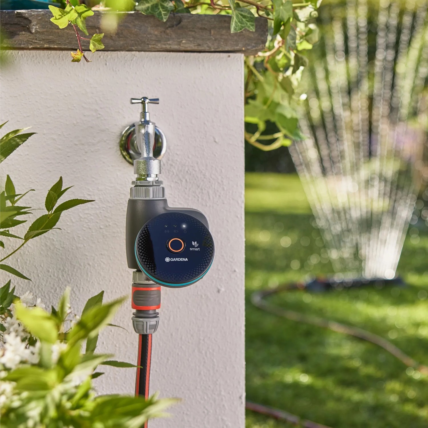 Gardena Smart Water Control Bewässerungscomputer am Wasserhahn, Rasensprenger im Hintergrund.
