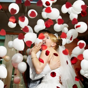 Glückliches Brautpaar feiert unter rotem Herzkonfetti und weißen Ballons. Konfettikanone für Hochzeit & Party.