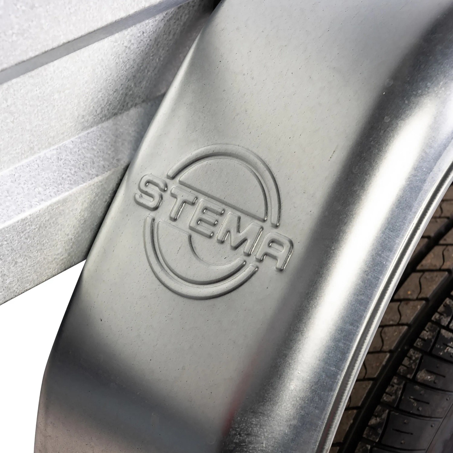 Detailaufnahme: Silberner Stema Autoanhänger Kotflügel mit Reifen und Logo.