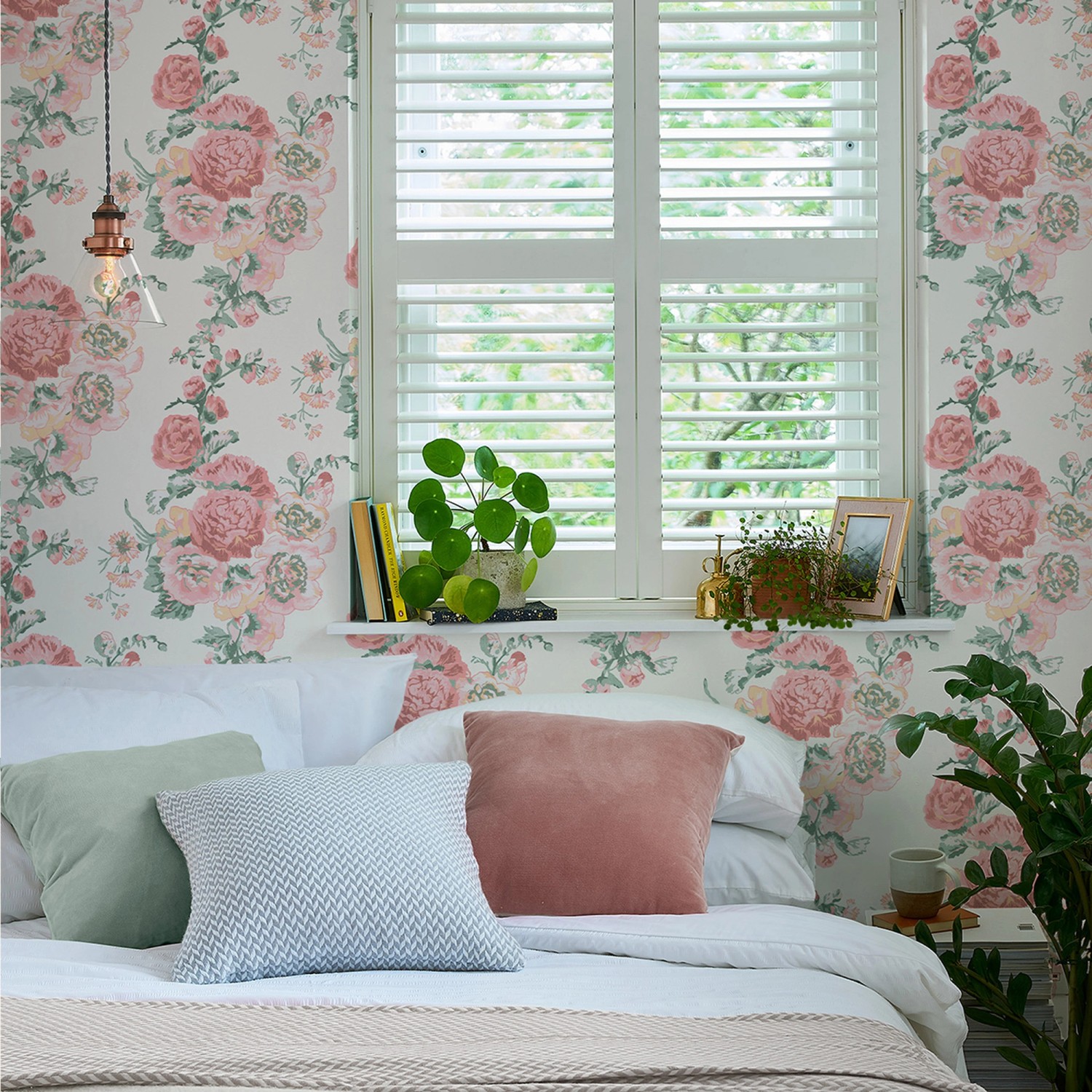 Laura Ashley Vliestapete Hollyhocks in Rosa/Koralle im Schlafzimmer mit Kissen und Pflanzen.